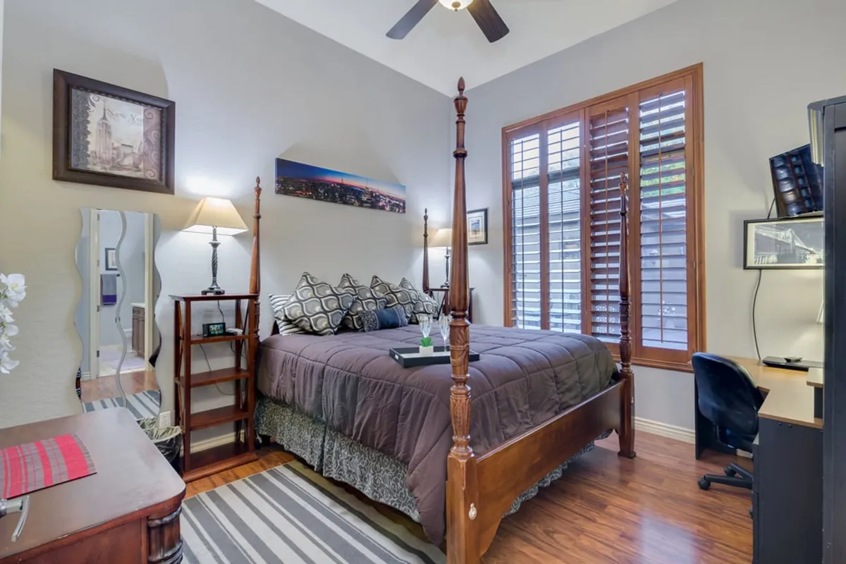 A serene bedroom featuring a four-poster bed with decorative pillows, a desk with chair, soft lamplight, and artistic wall decor. The presence of a workspace within the bedroom signals versatility for both relaxation and productivity.