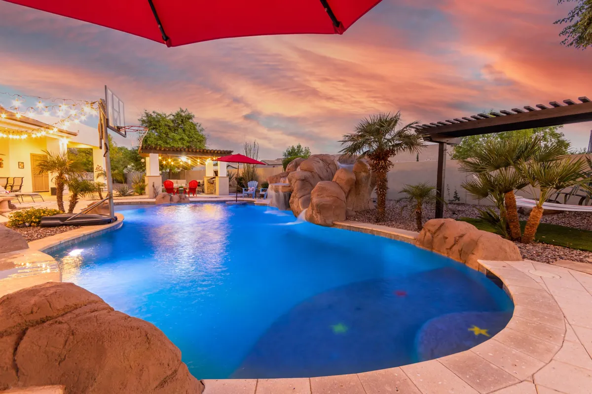 A wide-angle view of a blue swimming pool accented by natural rock formations, red umbrellas, palm trees, and an outdoor seating area. The space feels expansive, private, and luxurious, with playful and relaxing elements seamlessly combined.