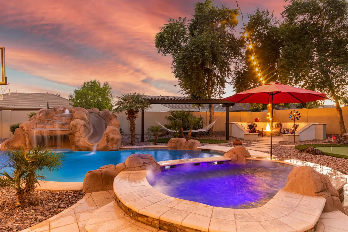Drawing the eye to a beautiful poolside scene, this image features a waterfall, a shaded seating area with an umbrella, a hammock, and a cozy seating area, all set against lush landscaping. The overall ambiance is serene and inviting, perfect for both relaxation and group gatherings.