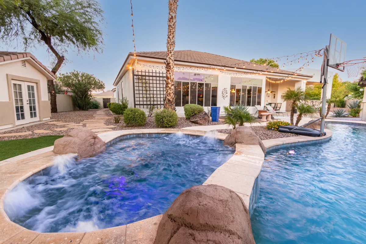 A vibrant backyard scene featuring a swimming pool, seating area, palm trees, decorative lighting, and a basketball hoop. The area is perfect for swimming, barbecuing, playing basketball, or relaxing with friends and family in a joyful setting.