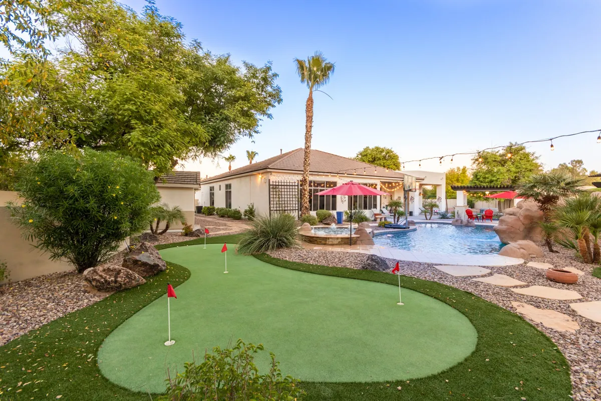 The image showcases the home's private backyard oasis at sunset, featuring a sparkling heated swimming pool surrounded by lush palm trees and vibrant landscaping. A modern patio with shaded umbrellas invites outdoor dining and relaxation, while a custom putting green offers fun for all ages. The atmosphere is serene yet lively, ideal for both unwinding and entertaining.
