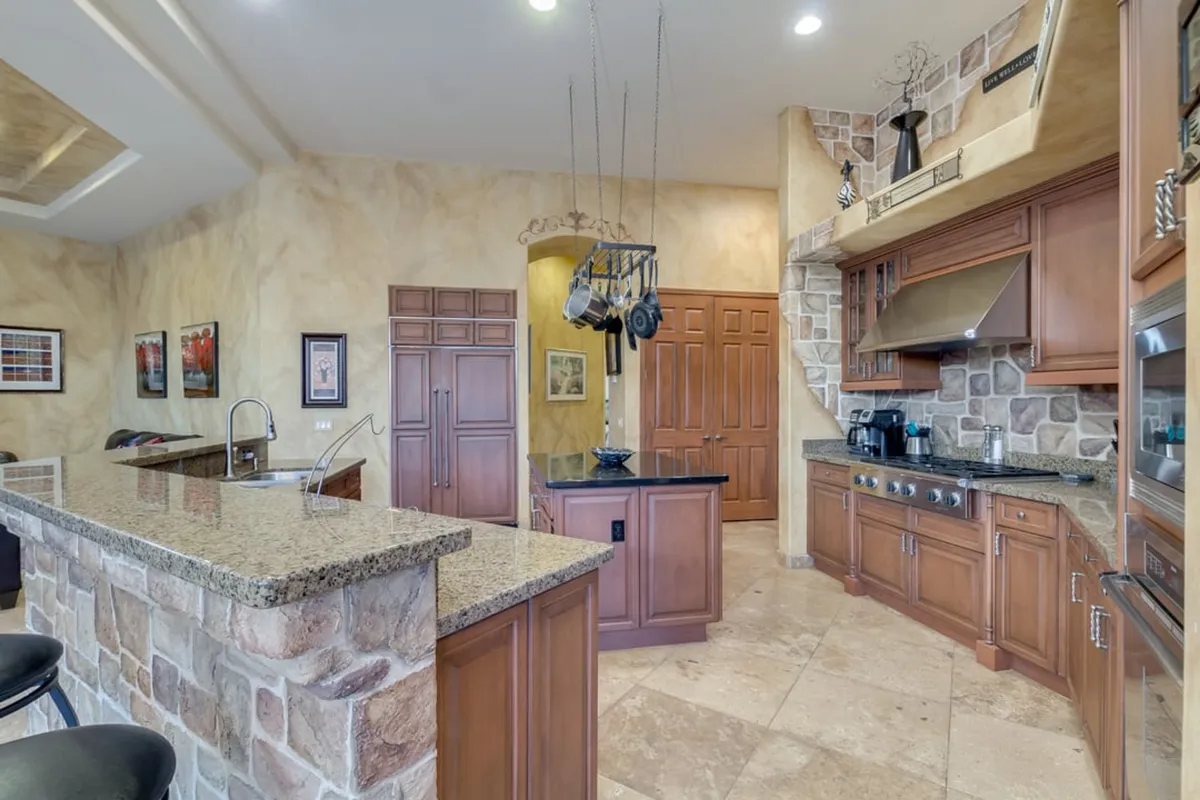 The kitchen showcases a spacious island with granite countertops, hanging pots, a stone backsplash, and rich wood cabinetry. The layout is open and elegant, facilitating both cooking and social interaction.