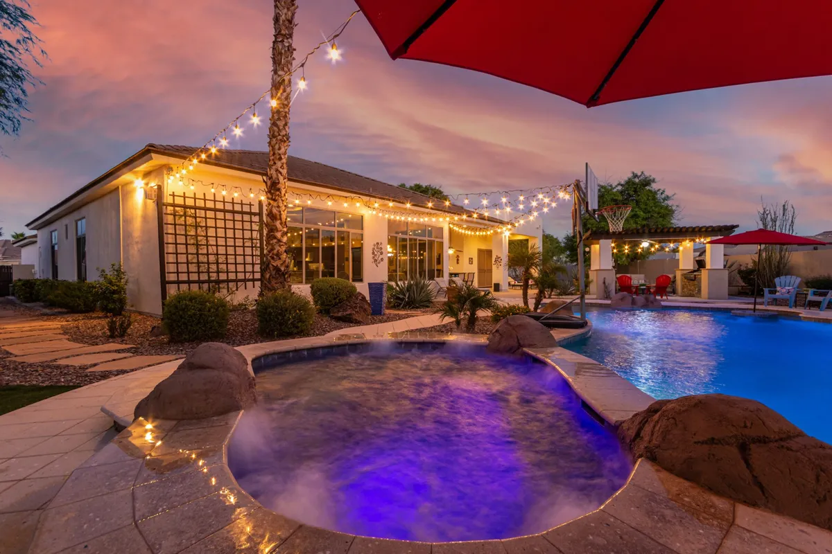 A vibrant exterior view of the backyard showcases a swimming pool, hot tub, string lights, a red umbrella, and a beautifully landscaped garden. The space is inviting for both relaxation and social gatherings, highlighting amenities that make remote work enjoyable.