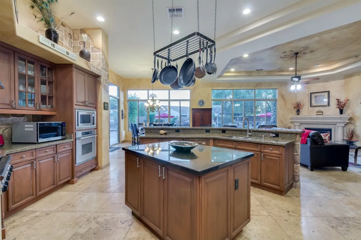 A kitchen and living space blend together, featuring a central island with a dark countertop, hanging cookware, abundant natural light, and a fireplace with decorative accents. The space is designed for both culinary creativity and comfortable relaxation.