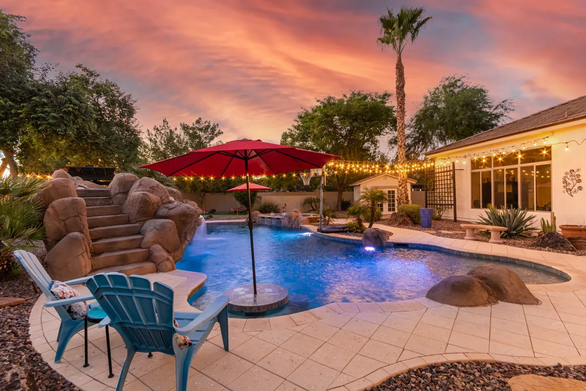 This image captures a vibrant backyard scene featuring a sparkling swimming pool with rock steps, a bold red umbrella for shade, string lights, and palm trees in the background. The inviting atmosphere suggests both daytime fun and relaxed evening gatherings, connecting directly to the description of a resort-style backyard with a heated pool and rock slide.
