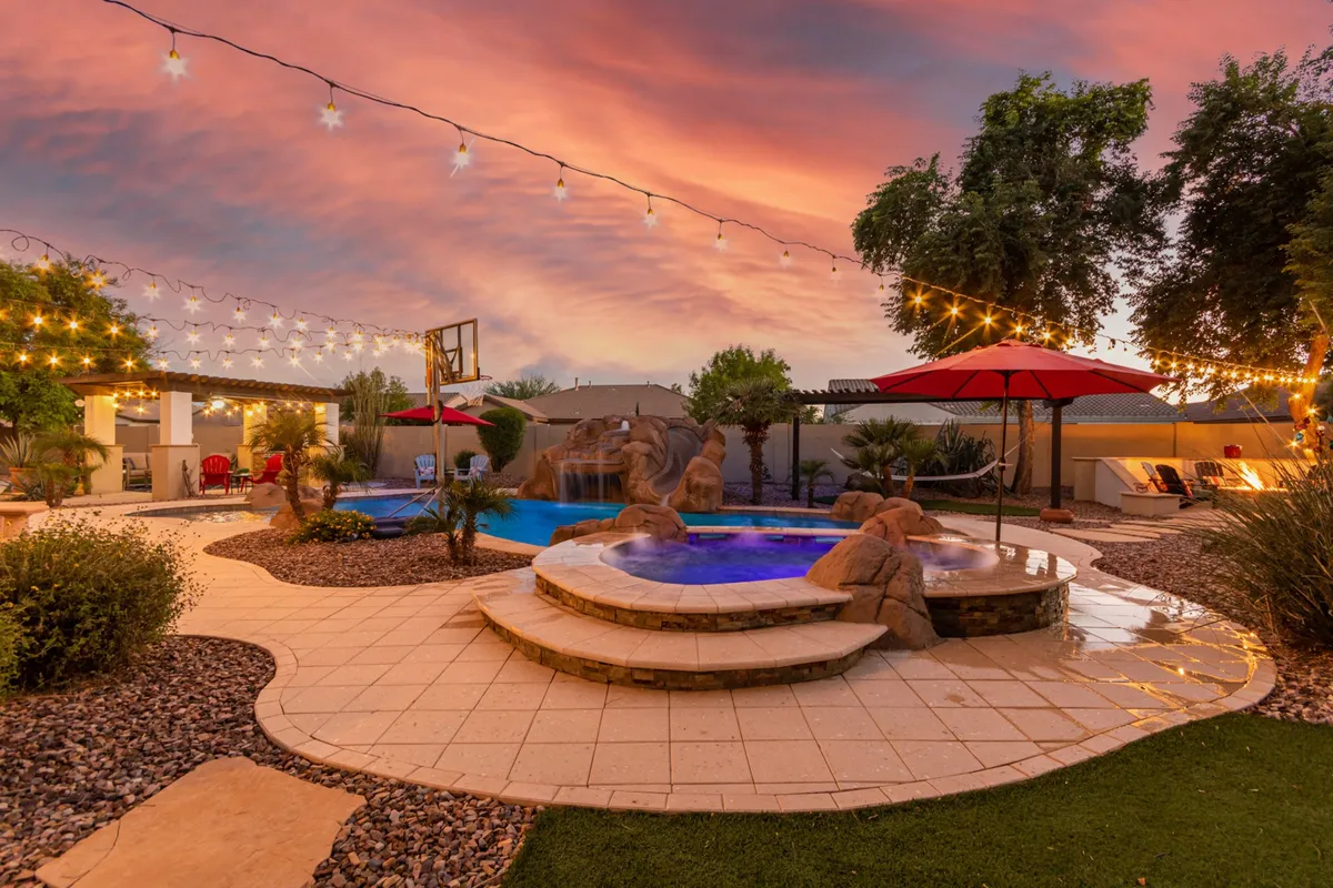 A stunning backyard view featuring a sparkling pool with a cascading waterfall, a spacious hot tub, and festive string lights overhead. The patio area and basketball hoop add to the lively, resort-like setting, ideal for evening gatherings and year-round enjoyment.