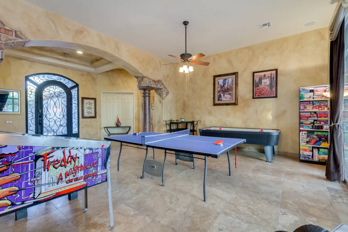 A well-lit, energetic entertainment area featuring a ping pong table and an arcade game machine. Game shelves are filled with board games, and decorative paintings line the walls. The arched doorways and open layout make the space feel inviting and inclusive for all ages.