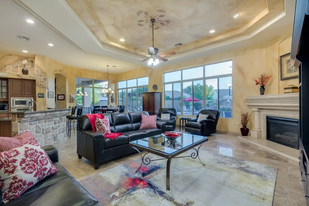 This image captures a welcoming open-concept living area where a plush sofa and coffee table face a fireplace, with large windows illuminating the space and a kitchen area visible in the background. The design is both inviting and functional, ideal for gatherings and shared experiences.