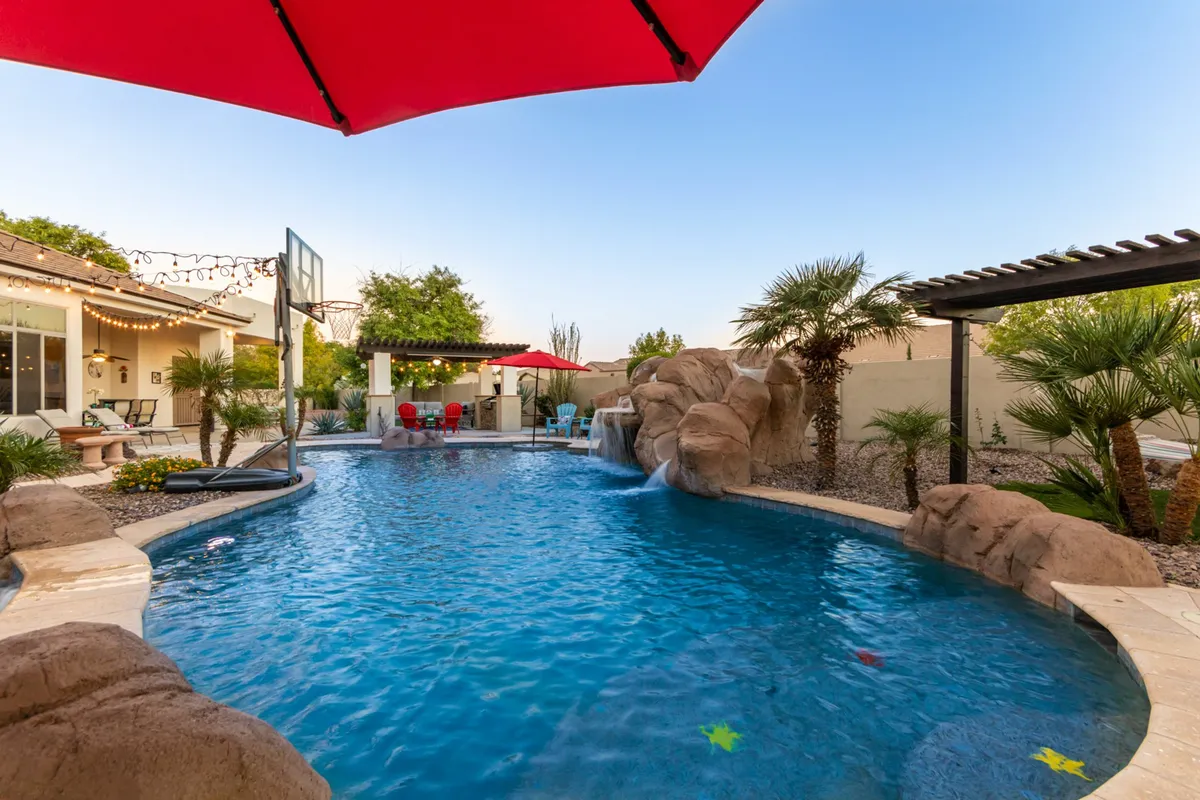 This image showcases a vibrant swimming pool surrounded by lounge chairs and a red umbrella, with a striking waterfall feature and a basketball hoop visible. The landscaping around the pool enhances the luxurious atmosphere, and the overall scene suggests both relaxation and playful energy.