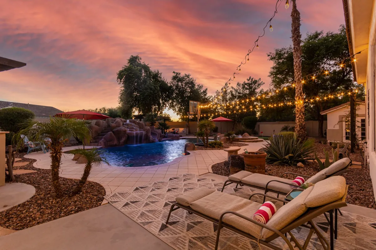 This image captures a serene backyard scene featuring a sparkling swimming pool surrounded by lush landscaping, poolside loungers, string lights, and a beautiful sunset sky. The tranquil setting is perfect for relaxation or entertaining between work sessions.