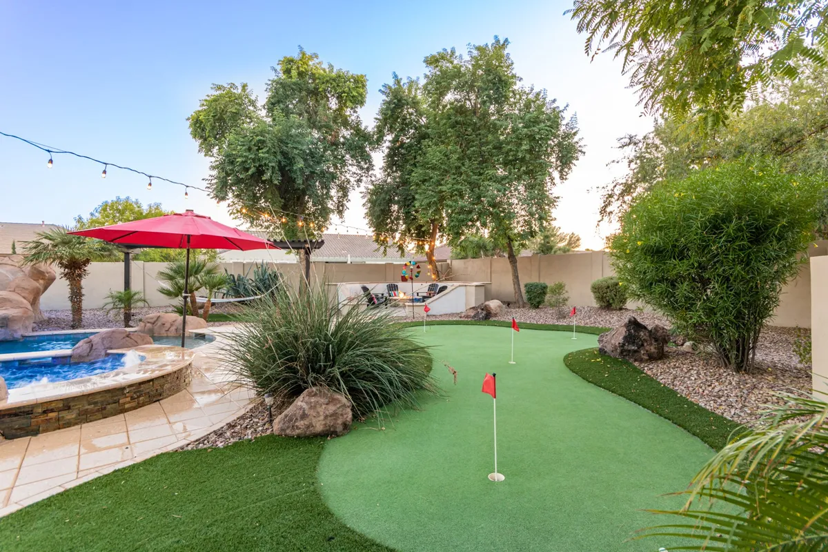 This exterior shot showcases a lushly landscaped yard with a putting green, a red umbrella providing shade, and a pool area surrounded by rocks and greenery. It conveys an active, playful mood, ideal for children and adults to enjoy friendly competitions or leisurely afternoons.